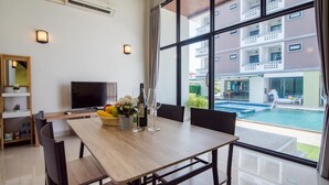 A modern hotel room with a dining table, chairs, a TV, and a large window overlooking a swimming pool and building. - Goodtimes Pool Villa 2 (Walk TO Beach) (Hua Hin)