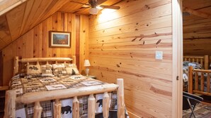 A cozy cabin bedroom with a wooden bed, a ceiling fan, and a painting on the wall. -  Rustic Log Lodge Overlooking Water and Mountain Views (Stanley)