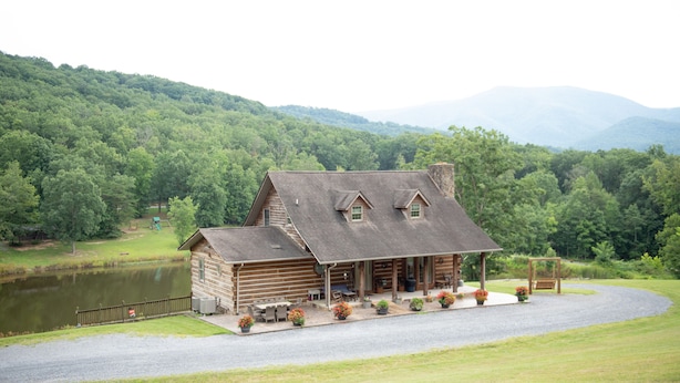 A log cabin with a porch, a pond, and a mountain backdrop. -  Rustic Log Lodge Overlooking Water and Mountain Views (Stanley)
