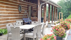 A wooden cabin with a covered patio area featuring wicker furniture and potted plants. -  Rustic Log Lodge Overlooking Water and Mountain Views (Stanley)