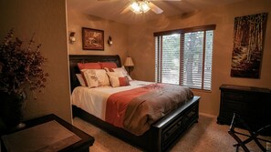 Une chambre à coucher comprenant un lit, une table de chevet, une commode, une fenêtre avec des stores et un ventilateur de plafond.