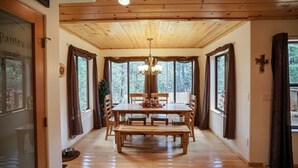 Uma área de jantar com mesa e cadeiras de madeira, um lustre e janelas amplas com cortinas.