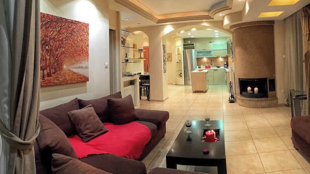 A living room with a sofa, a coffee table, and a fireplace. There is a kitchen in the background with a refrigerator and cabinets. - Elegant Vouliagmeni apartment in a Unique Spot. (Vouliagmeni)