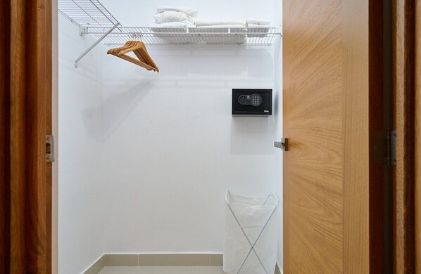 Une chambre d’hôtel avec une porte en bois, un mur blanc, une étagère en métal avec des serviettes, un clavier de sécurité et un vêtement suspendu.