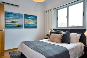 Un dormitorio con una cama, dos cuadros, una ventana y una mesita de noche con lámpara.