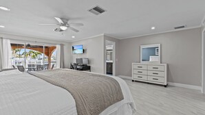 A spacious bedroom with a large bed, a ceiling fan, a TV, a dresser, and a view of the outdoors. - Water Front Home with Heated Pool & Hot Tub, 2 Master Suites & a Floating Dock (New Port Richey)
