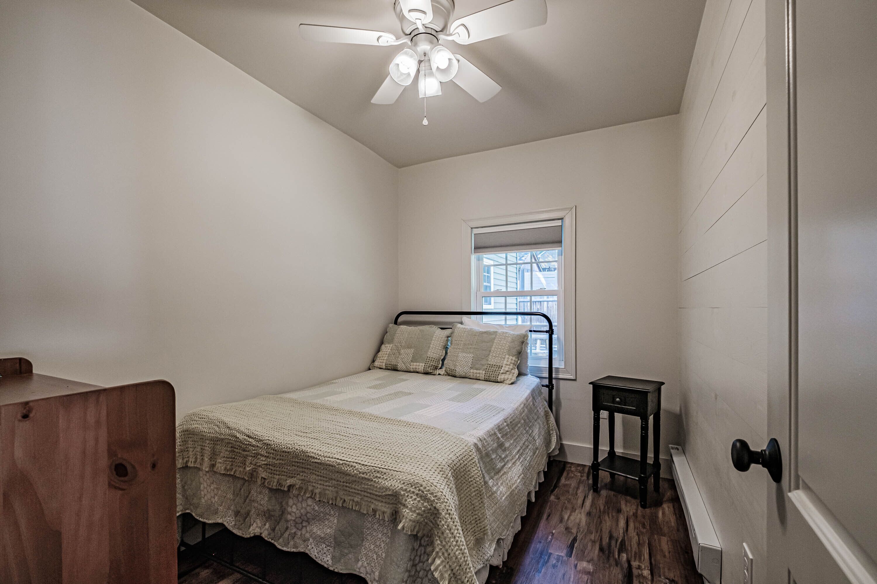 A bedroom with a bed, a ceiling fan, a window, and a small table.
