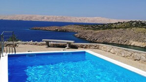A swimming pool with clear blue water, a stone bench, and a view of the sea and rocky hills in the background. - Villa Mihaela - Beachfront apartment "Ika" with sea view and pool (Cesarica)