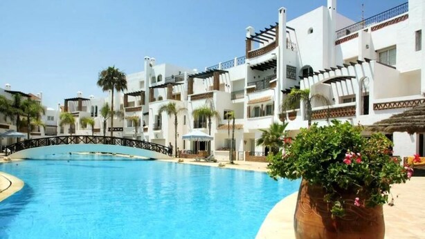 Une piscine entourée de bâtiments blancs dotés de balcons, de palmiers et d’un grand pot de fleurs contenant une plante aux fleurs roses.