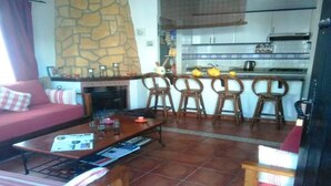 Un salon comprenant un canapé, une table basse et des fauteuils, un mur en pierre et un coin cuisine avec un comptoir et des chaises.