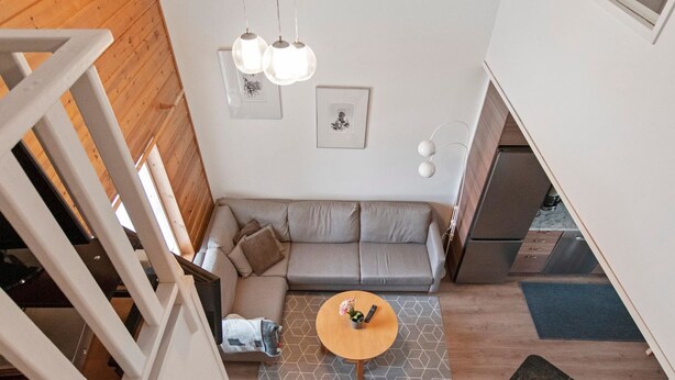 Ein moderner Loft-Wohnraum mit einem grauen Ecksofa, einem Holztisch und einer Treppe, die zu einer oberen Ebene führt.