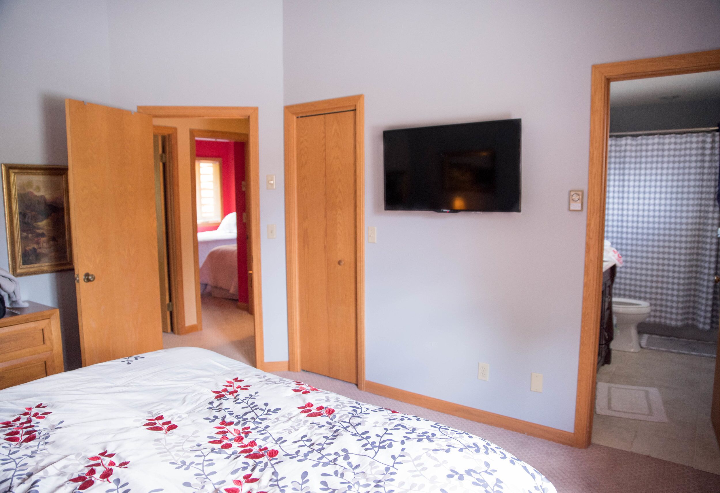 A bedroom with a bed, a TV mounted on the wall, and a bathroom visible through an open door.