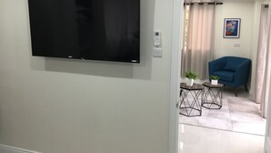 A living room with a flat-screen TV mounted on the wall, a blue armchair, a small table with a plant, and a painting on the wall.