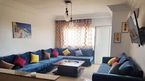Een woonkamer met een blauwe zithoek, een salontafel en een aan de muur bevestigde televisie.