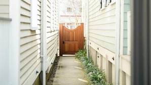 Een smal betonnen pad, begrensd door witte gebouwen met groene accenten, en een houten poort aan het einde.