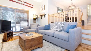 A living room with a grey sofa, wooden coffee table, and a TV stand. There is a staircase with wooden railings and a chandelier. - Ski to the door!True SI/SO Luxury 2BrTH-Private HT (Whistler)