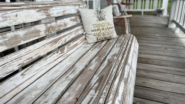 Un banc en bois usé par les intempéries, agrémenté d’un coussin sur lequel est brodé le message : « Tenez-vous droite, portez une couronne et soyez toujours gentille. »