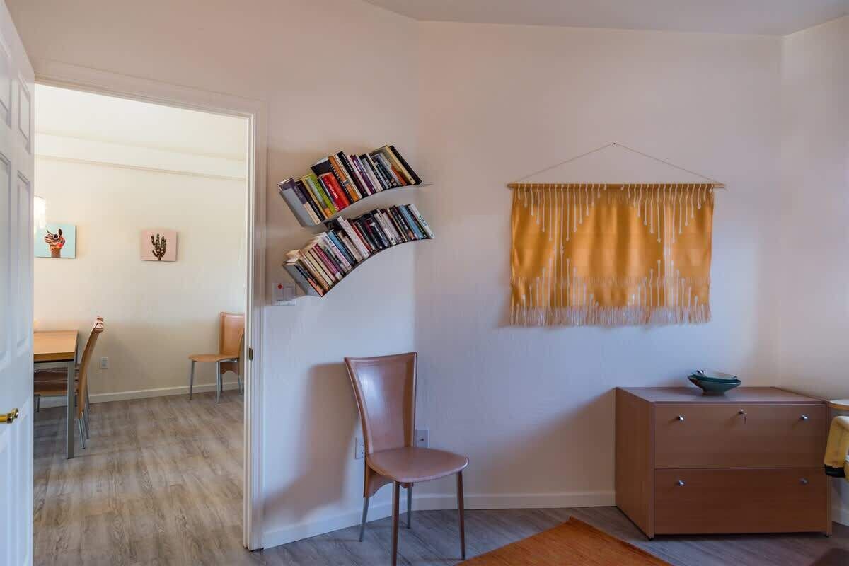A room with a curved bookshelf, a single chair, a small cabinet, and a window with a yellow curtain.