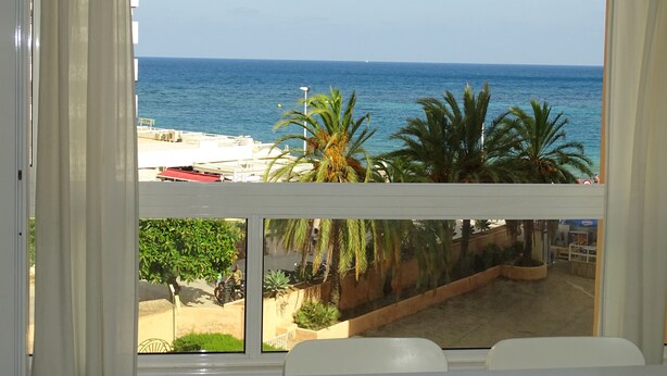 Une vue depuis une fenêtre donnant sur une plage bordée de palmiers, sous un ciel dégagé.