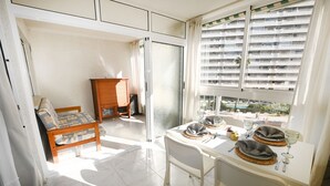 Un coin repas avec une table dressée pour deux personnes, un banc en bois et une vue sur un immeuble d’habitation à travers une fenêtre munie de stores.