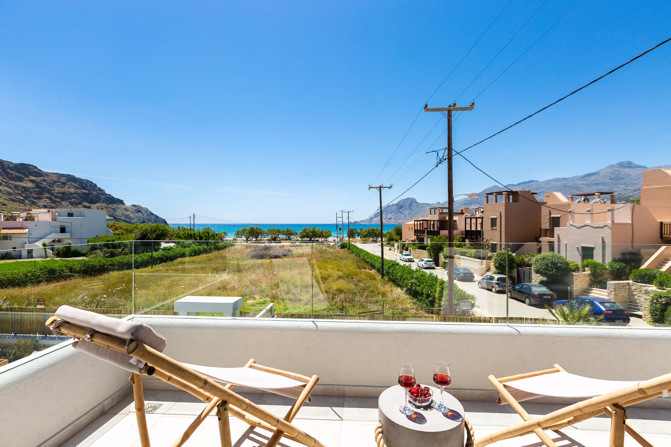 A balcony with two chairs, a table with two glasses of red wine, and a view of a coastal street with cars and buildings.