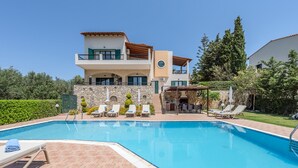 Una casa de dos pisos con piscina, mobiliario exterior y un muro de piedra.