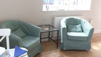 Une pièce avec deux fauteuils verts, une petite table en verre, un radiateur blanc et une fenêtre donnant sur des immeubles.