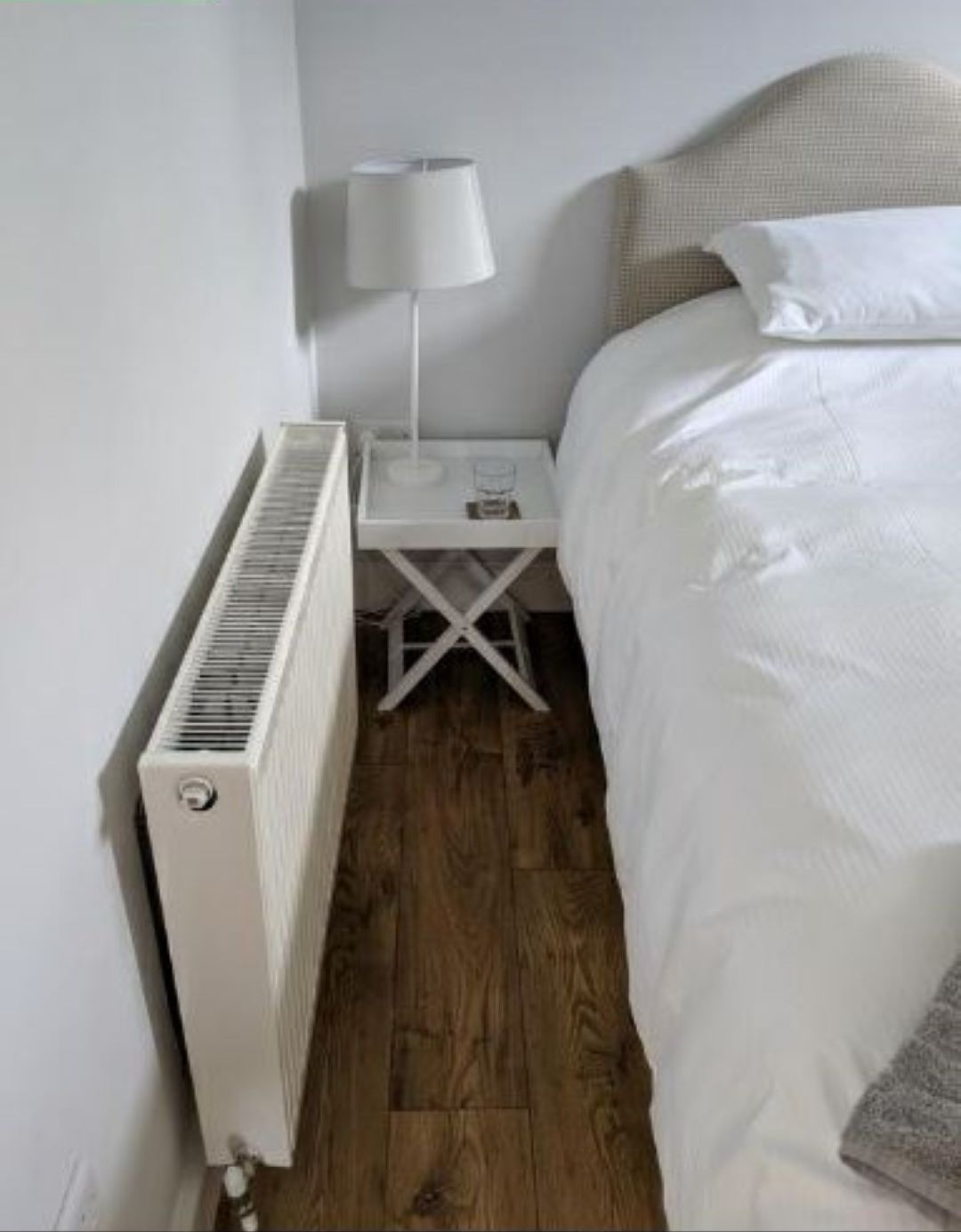 Een slaapkamer met een witte radiator, een nachtkastje met een lamp, en een bed met witte lakens.