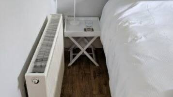 Een slaapkamer met een witte radiator, een nachtkastje met een lamp, en een bed met witte lakens.