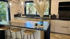 A modern kitchen with a central island, bar stools, pendant lights, and a view of greenery outside. - New villa 185m2 located close to beaches, port and city center (Porto-Vecchio)