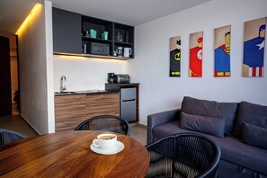 Eine moderne Küche mit Mikrowelle, Toaster und Kaffeemaschine. Ein Esstisch mit einer Kaffeetasse und schwarzen Stühlen steht daneben. Im Wohnbereich befindet sich eine graue Couch mit einem Bild an der Wand.