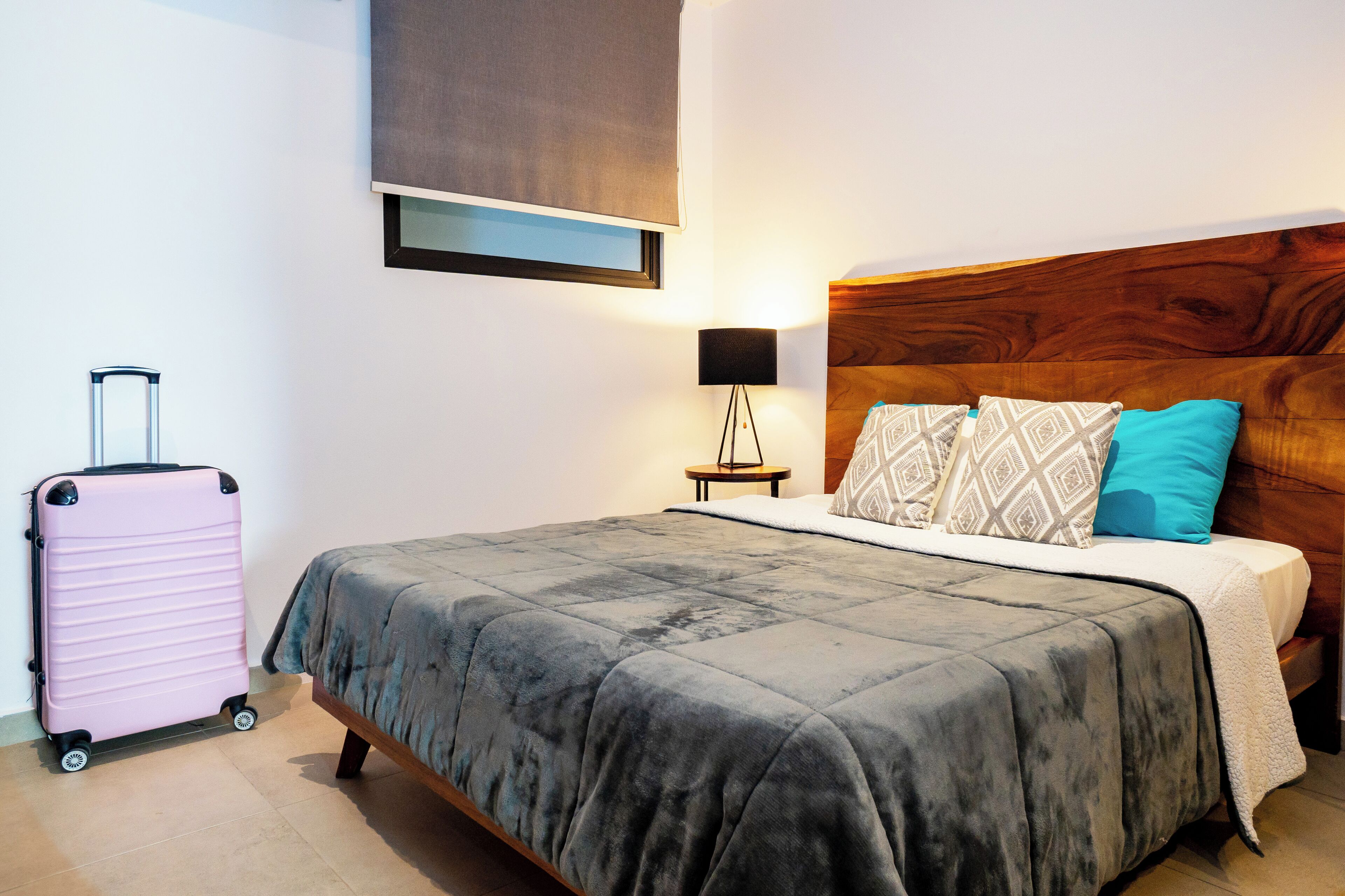 Ein ordentlich bezogenes Bett mit einer grauen Bettdecke, einem Holz-Kopfteil und zwei Kissen mit einem geometrischen Muster sowie einem schlichten blauen Kissen. Ein pinkfarbener Koffer steht an der Wand neben dem Bett.