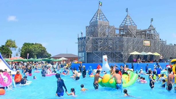Una piscina concurrida, gente disfrutando del agua, una gran estructura inflable y un edificio con un diseño arquitectónico llamativo al fondo.