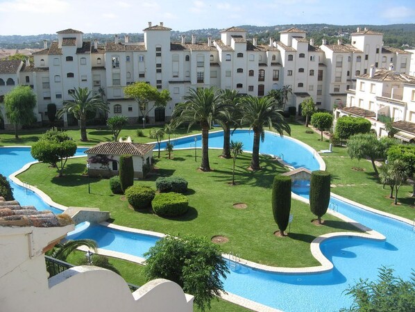 A resort with a large swimming pool, manicured lawns, and a row of white buildings with red-tiled roofs.