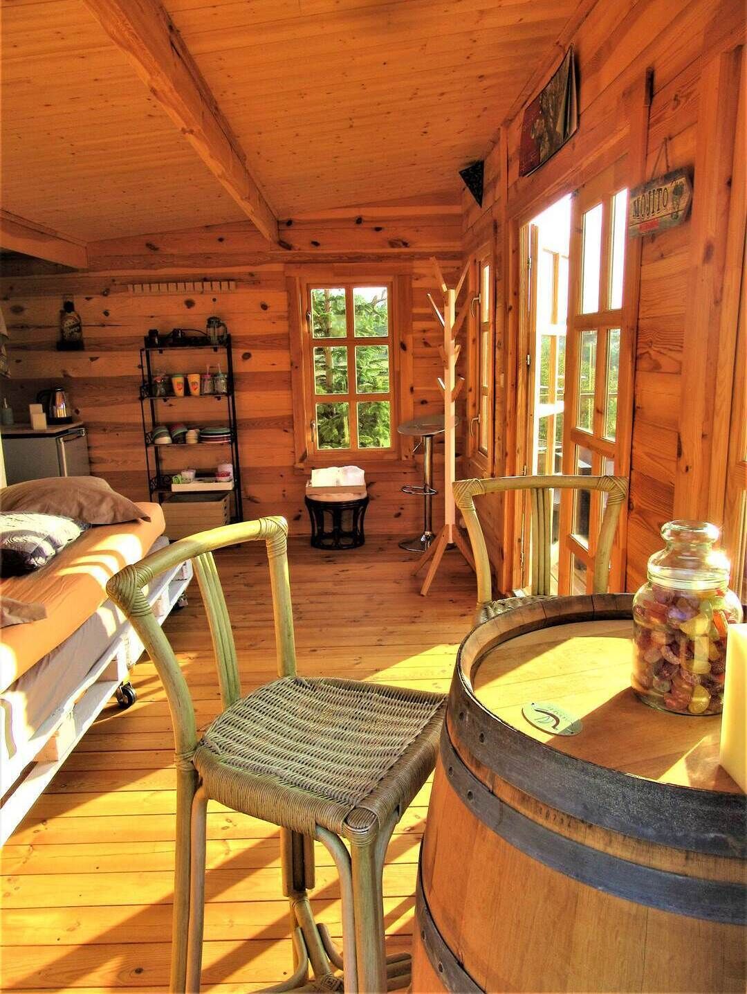 A rustic cabin interior with a wooden floor, a bed, a chair, a table, a barrel, and a window with a view of trees.