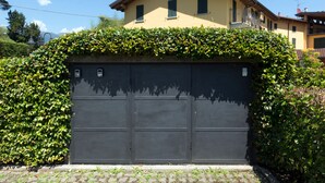 Une porte de garage avec une haie devant, un bâtiment jaune avec un toit brun en arrière-plan, et un ciel bleu dégagé.