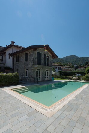 Uma moradia de pedra com piscina, rodeada por um pátio pavimentado e vegetação exuberante.