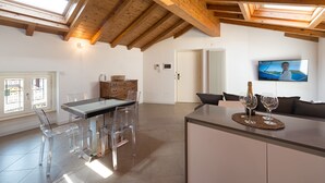 Une cuisine moderne avec un plafond en bois, un coin repas avec des chaises transparentes, un téléviseur fixé au mur et un coin salon confortable avec un canapé.