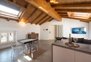 Une cuisine moderne avec un plafond en bois, un coin repas avec des chaises transparentes, un téléviseur fixé au mur et un coin salon confortable avec un canapé.