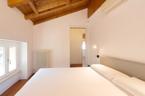 Quarto com teto de madeira, radiador, cama com roupa de cama branca e janela com vista.