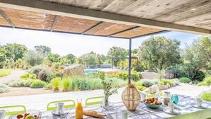 Un área de comedor al aire libre con una mesa lista para el desayuno, rodeada de vegetación y vistas a una piscina.