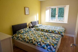 Two beds with geometric-patterned bedspreads, a window with a view of greenery, and a framed picture adorn this inviting space.