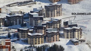 Ein schneebedecktes Skigebiet mit mehreren mehrstöckigen Gebäuden, vermutlich Unterkünften, und einer Seilbahn im Hintergrund.
