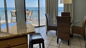 Een kamer met een keukentrap, een eethoek met rieten stoelen, een glazen tafel en uitzicht op de oceaan door grote ramen.