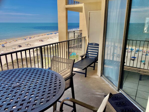 A balcony with a view of the beach, a table, and two chairs.