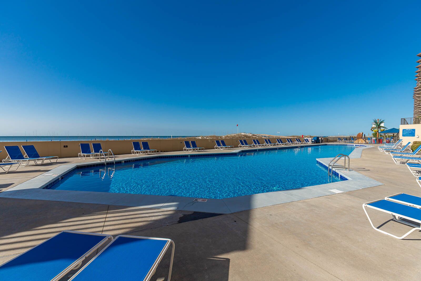 A pool area with blue lounge chairs and a clear view of the ocean.