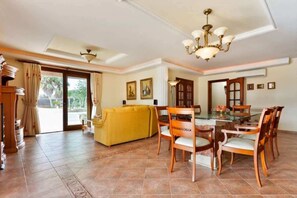 A spacious living room with a yellow sofa, wooden dining table, and chairs. There is a large window with curtains, a chandelier, and a view of the outdoors.