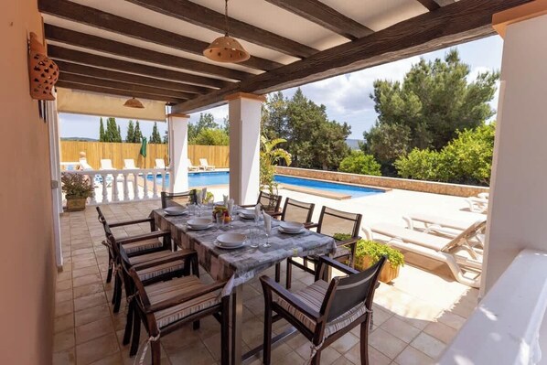 An outdoor patio with a dining table set for six, surrounded by chairs, overlooking a swimming pool and lush greenery.