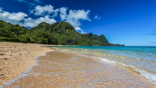 A beach with clear turquoise water, a sandy shore, and lush green hills in the background.