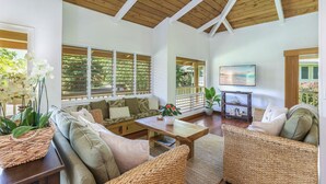 A spacious living area with a high ceiling, wooden beams, and large windows. There is a sofa set, a coffee table, and a TV on a stand.